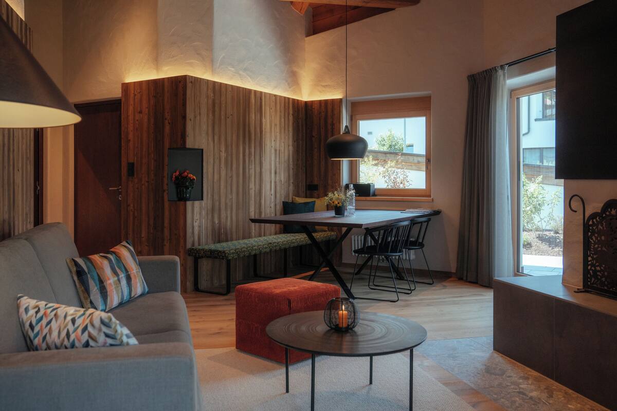 Un salon moderne avec un canapé, un pouf et une table basse. On y trouve également un coin repas avec une table et des chaises, et une cuisine visible au fond.