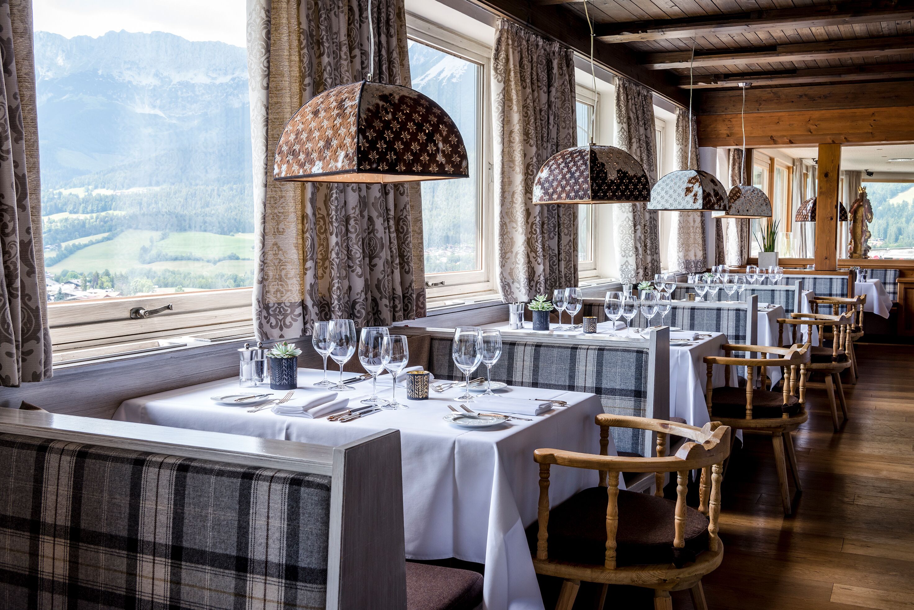 A restaurant with tables set for dining, large windows with mountain views, and wooden ceiling beams.