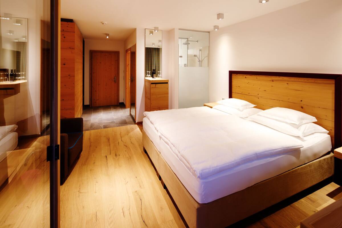 Une chambre d’hôtel moderne avec un plancher en bois, un grand lit avec du linge de lit blanc, une tête de lit en bois, une armoire en bois et une salle de bain avec une douche et un miroir.