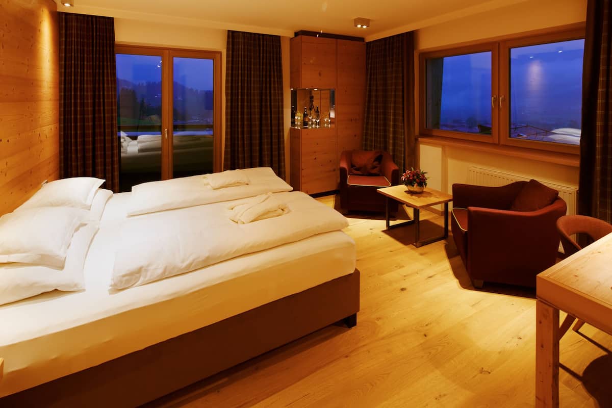 Une chambre d’hôtel avec un grand lit, deux fauteuils, une petite table et une vue sur l’extérieur grâce à de grandes fenêtres.