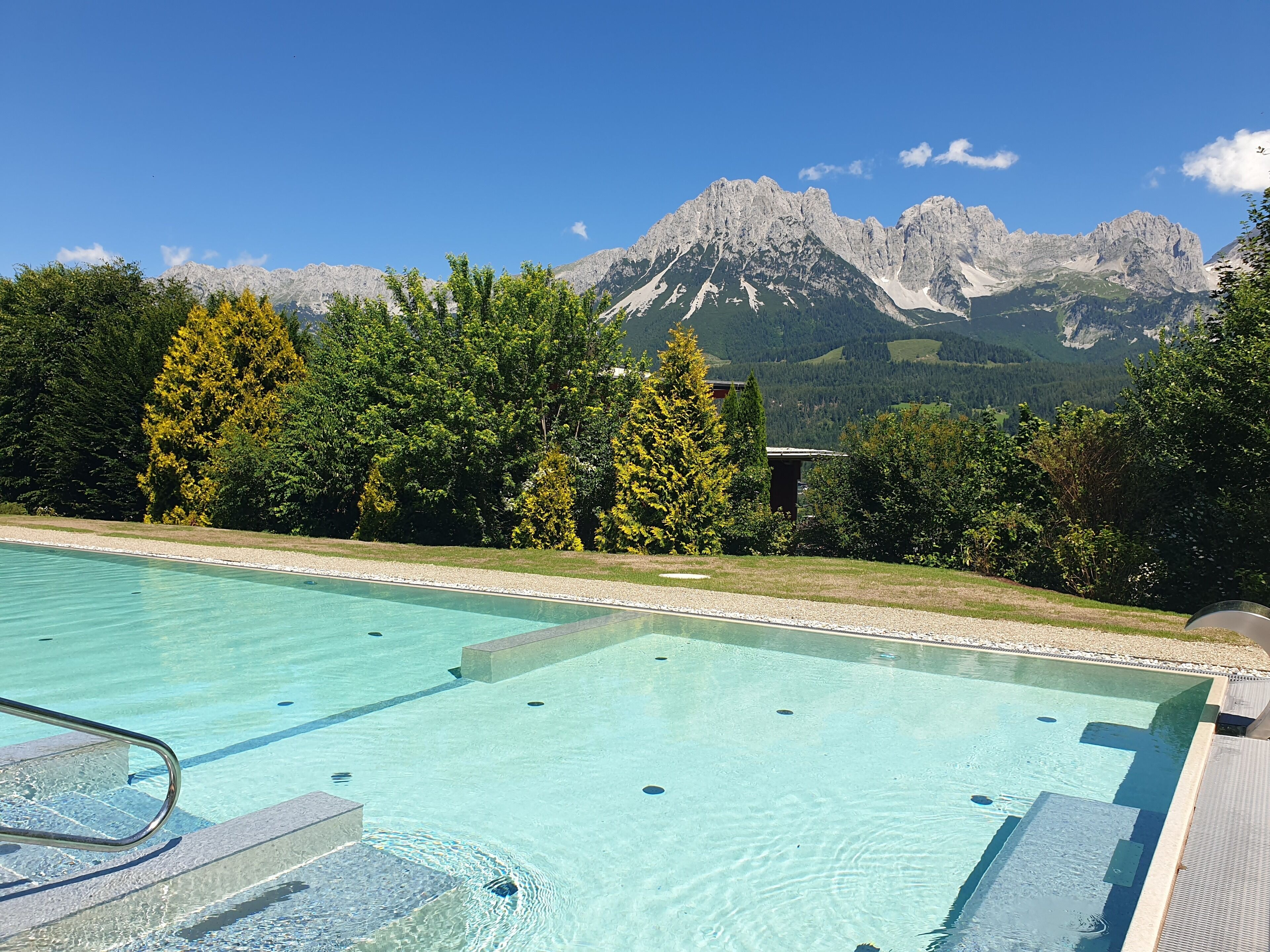Ein Schwimmbecken mit klarem Wasser, umgeben von Bäumen und Bergen im Hintergrund.
