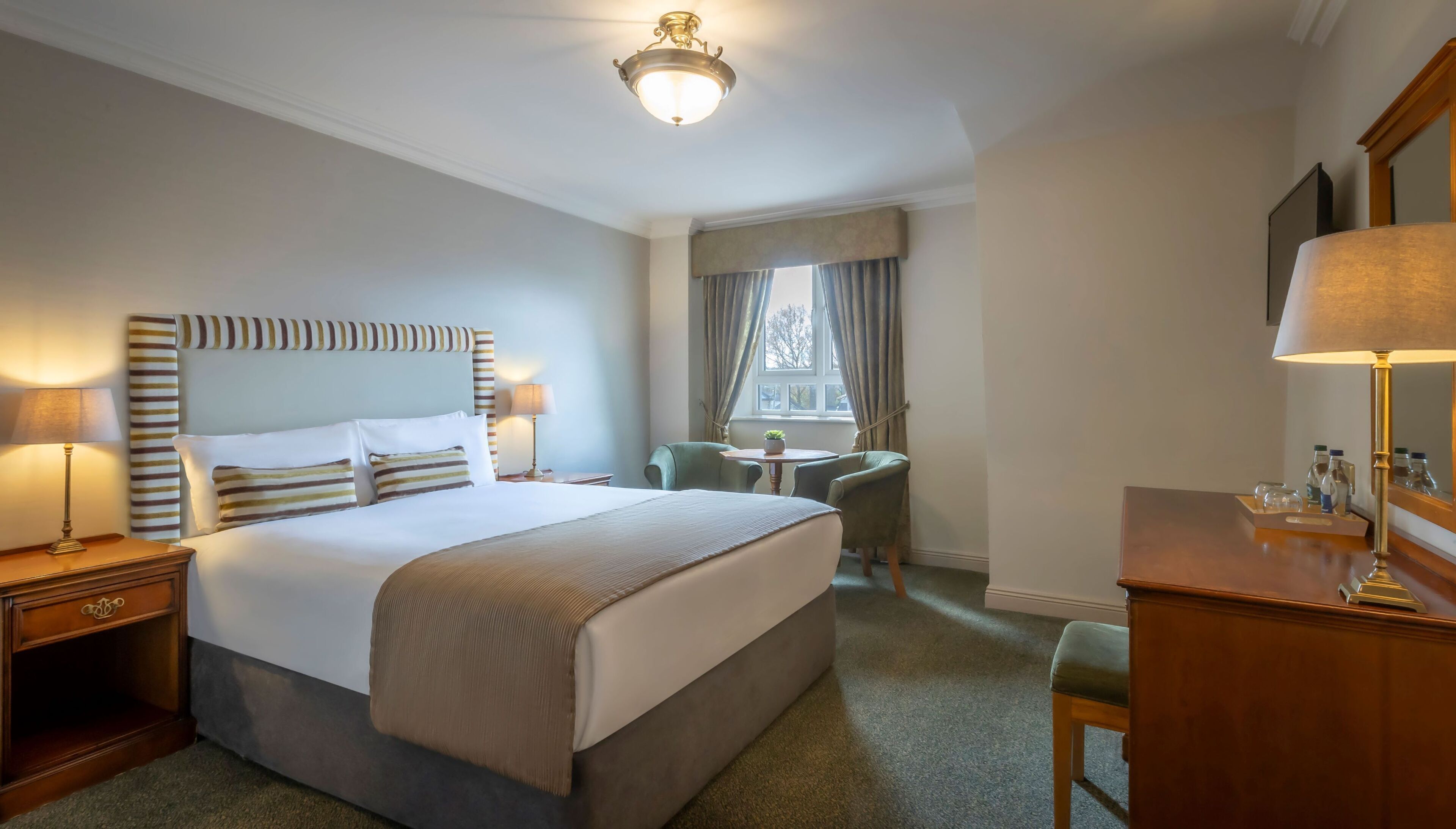 A hotel room with a large bed, bedside tables, a desk, a chair, a small table with a plant, and a window with curtains.