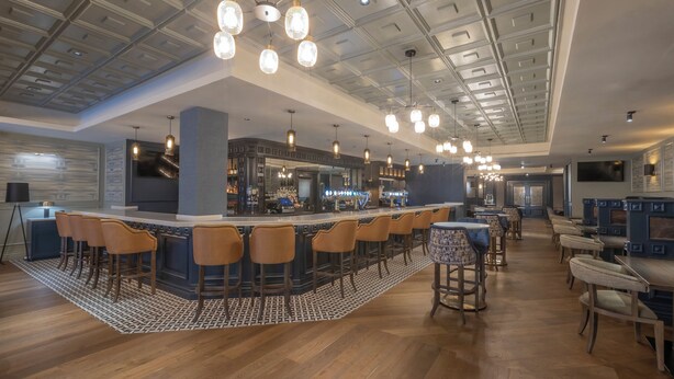 A modern bar area with a long counter, multiple stools, and hanging pendant lights. - Bracken Court Hotel (Balbriggan)