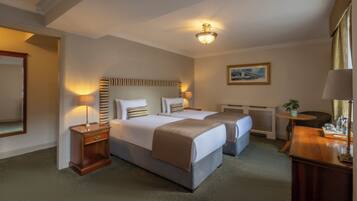 A hotel room with two beds, a nightstand, a desk, a chair, a painting, and a ceiling fan.