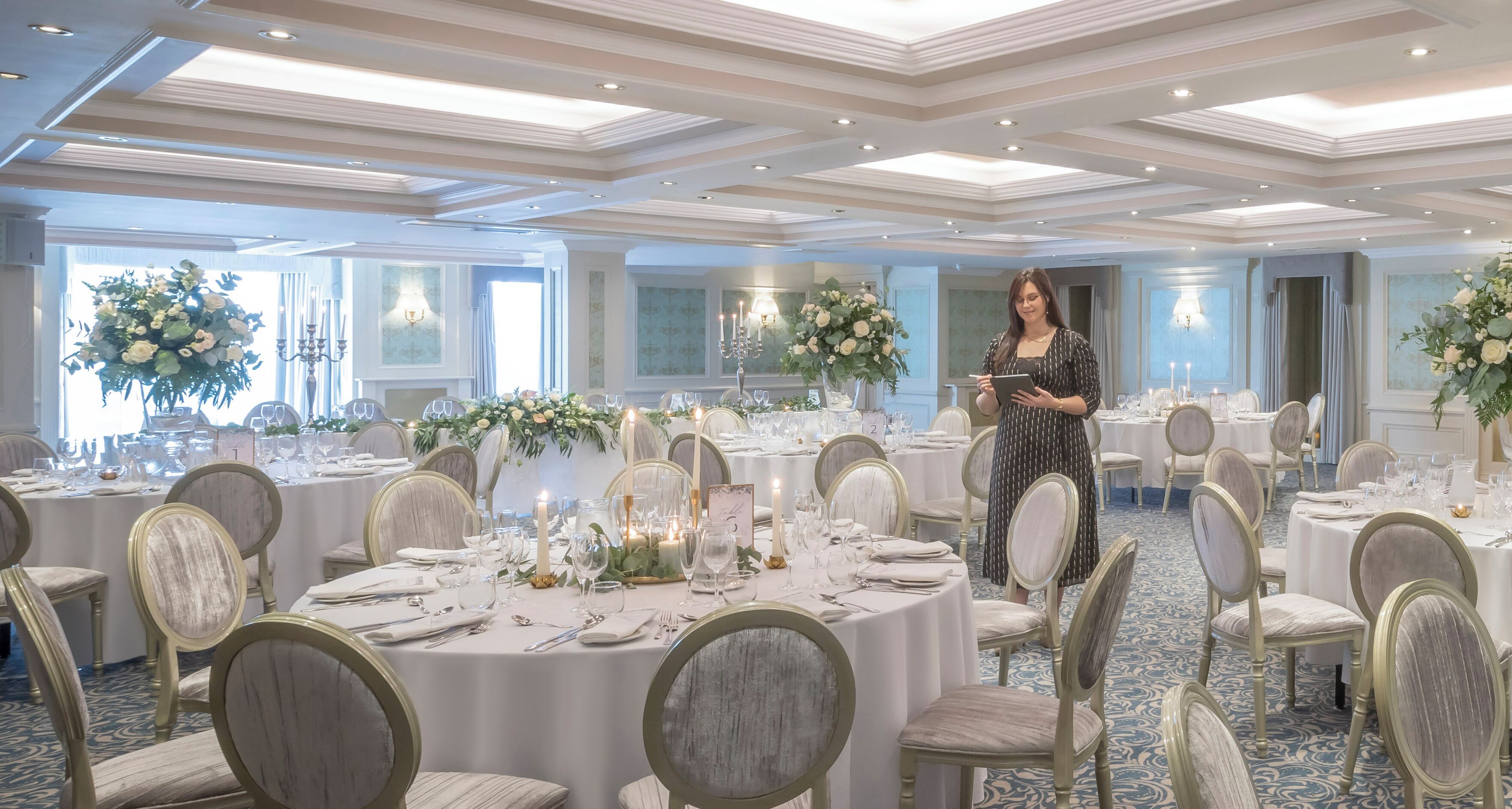 Un salón de banquetes con mesas redondas dispuestas para un evento formal, con candelabros, centros de mesa florales y suelo alfombrado.