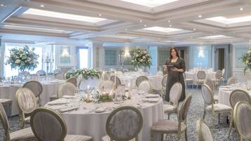 Un salón de banquetes con mesas redondas dispuestas para un evento formal, con candelabros, centros de mesa florales y suelo alfombrado.