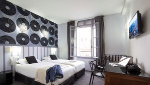 A hotel room with a bed, a desk with a laptop and camera, a TV, and a patterned wall. - Hôtel Raymond 4 Toulouse (Toulouse)