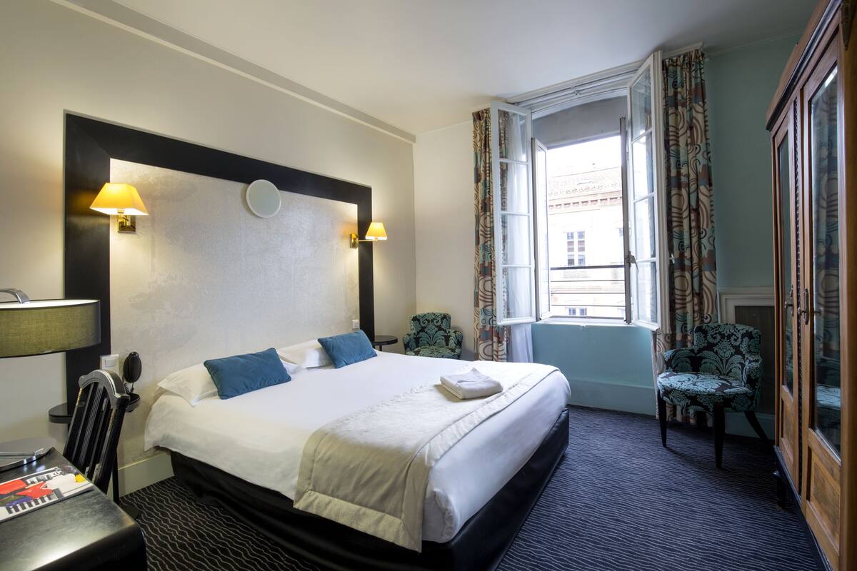 Een hotelkamer met een groot bed, twee bedlampen, een bureau met stoel, een raam met gordijnen en een kast.