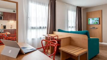 Una habitación de hotel moderna con un escritorio de madera, un sofá color turquesa, sillas rojas, una computadora portátil y un televisor montado en la pared.