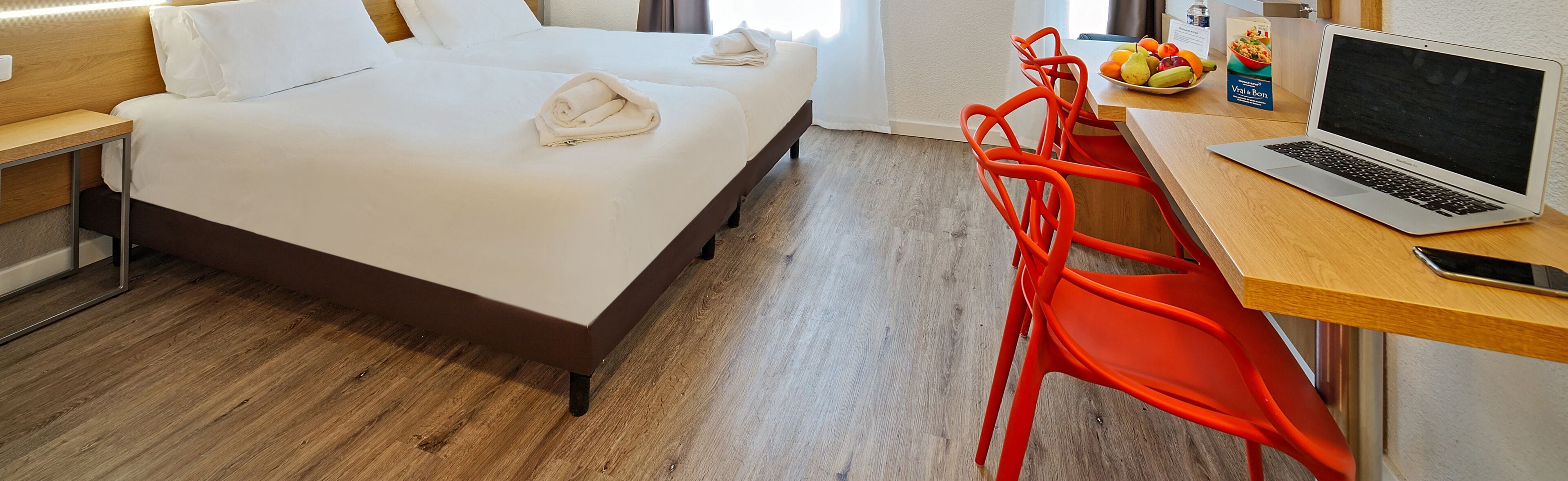 Een hotelkamer met een bed, een bureau met een laptop, een schaal fruit en een fles water.