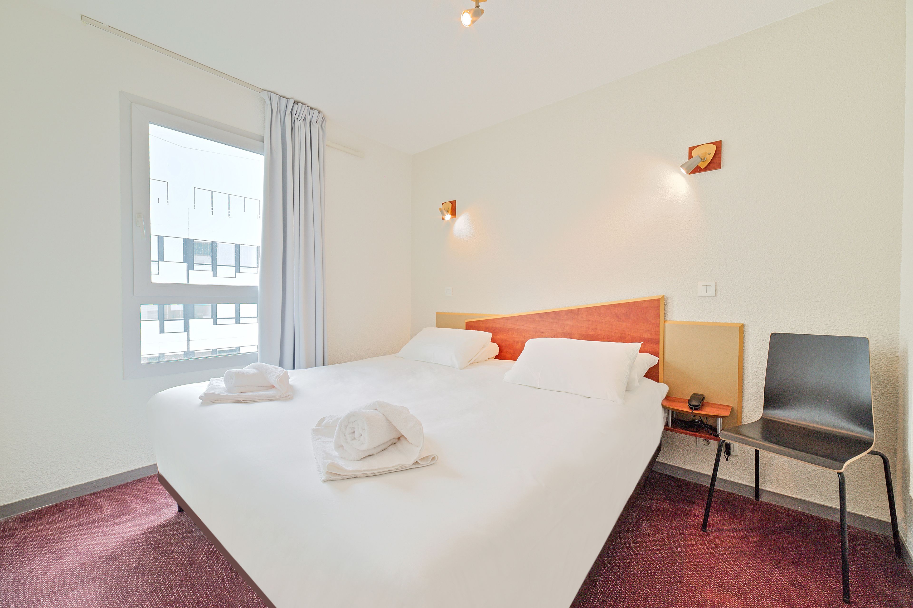 A hotel room with a large bed, a chair, a nightstand, and a window with curtains.