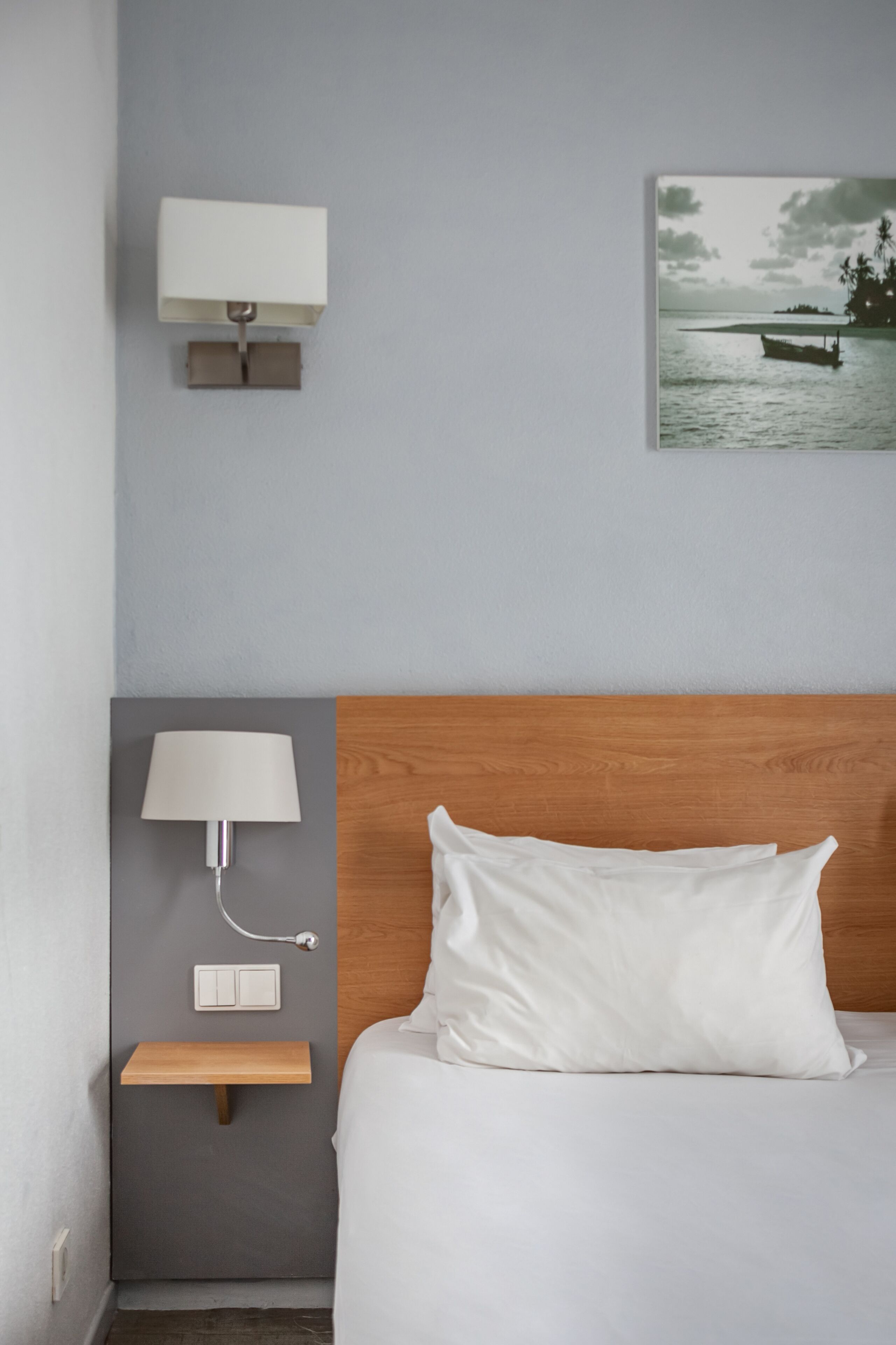 A neatly arranged hotel room with a wooden headboard, a white bed, a bedside table with a lamp, and a wall-mounted light fixture.