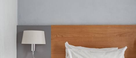 A neatly arranged hotel room with a wooden headboard, a white bed, a bedside table with a lamp, and a wall-mounted light fixture.