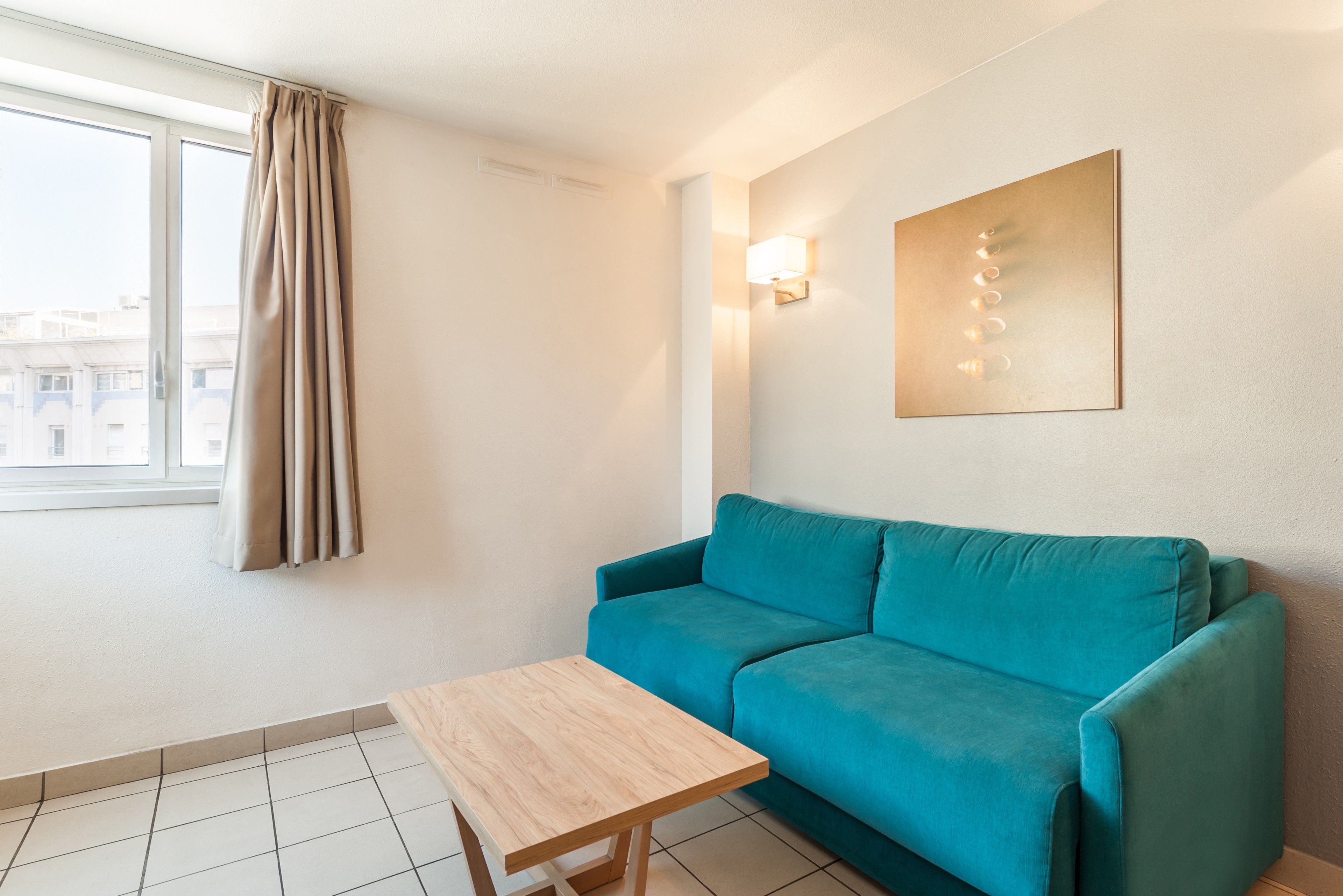 A room with a teal sofa, a wooden coffee table, a wall-mounted light, and a window with curtains.