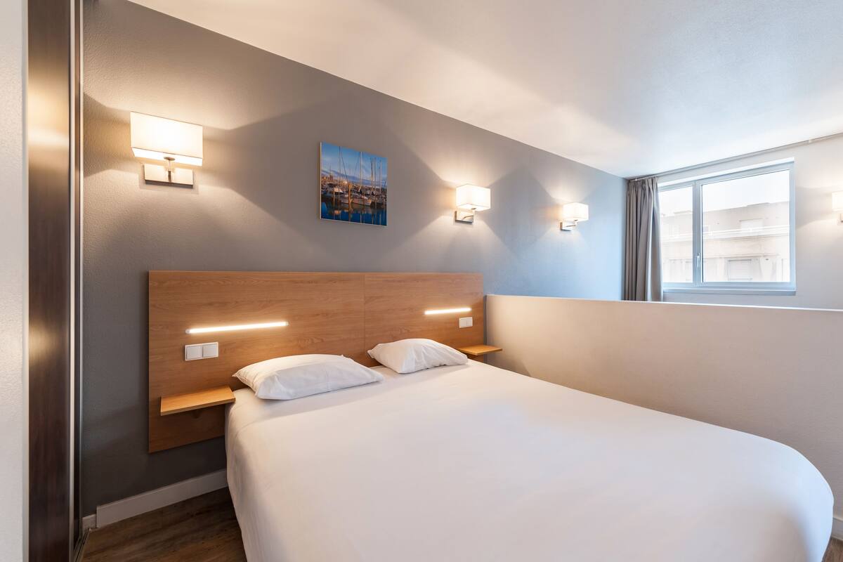 Ein Hotelzimmer mit einem großen Bett, zwei Nachttischen, Wandleuchten, einem Gemälde und einem Fenster mit Vorhängen.