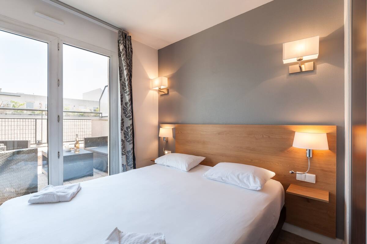 Ein Hotelzimmer mit einem großen Bett, zwei Nachttischlampen, einem Holzkopfende und einer Glastür, die auf einen Balkon mit Sitzmöbeln führt.