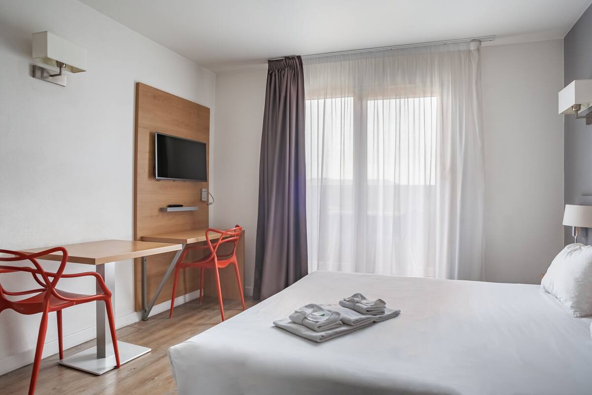 Ein Hotelzimmer mit Bett, Schreibtisch mit Stuhl, Fernseher und einem Fenster mit Vorhängen.