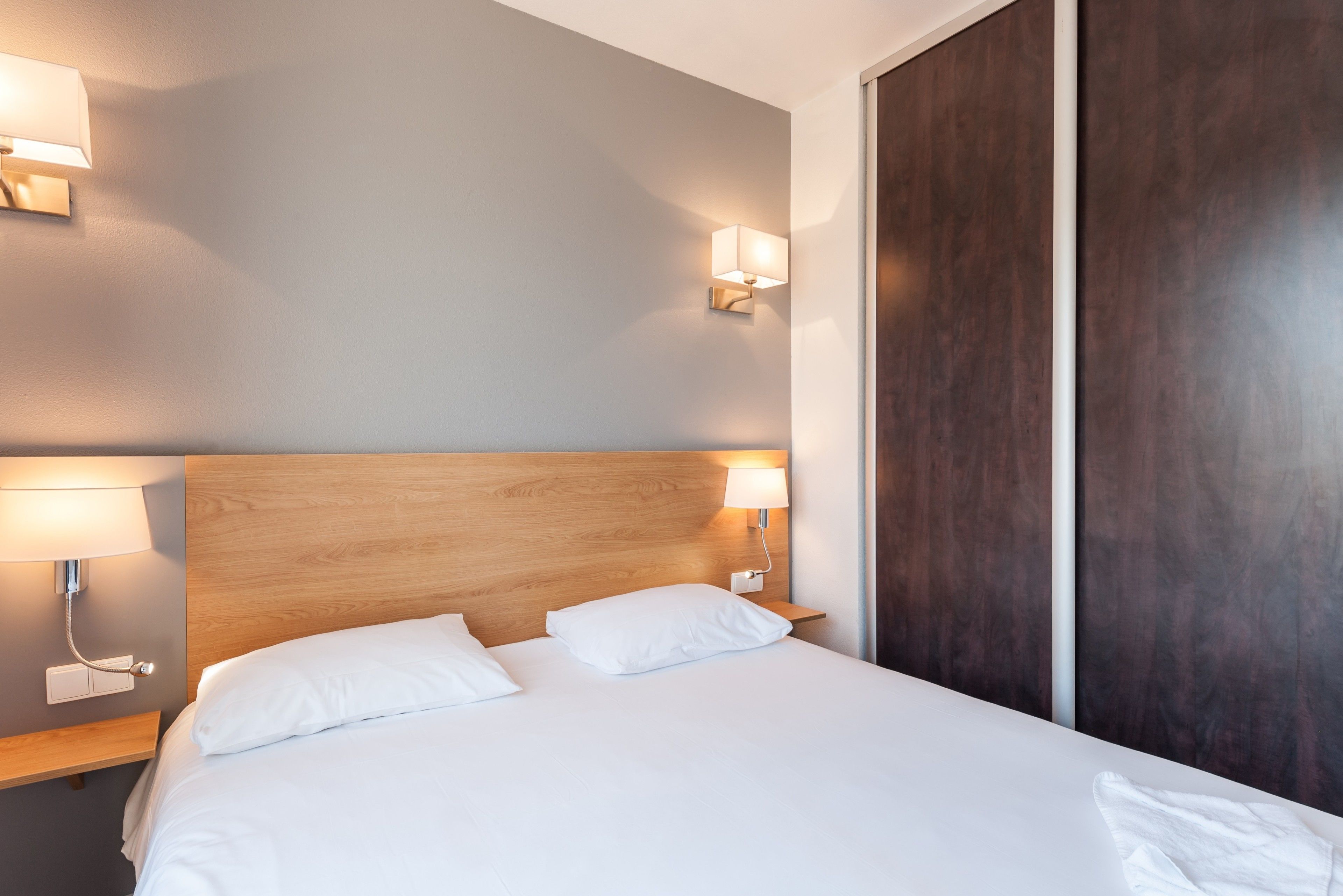 Habitación de hotel con cabecera de madera, dos lámparas de noche, una cama con ropa blanca y un armario de madera oscura.