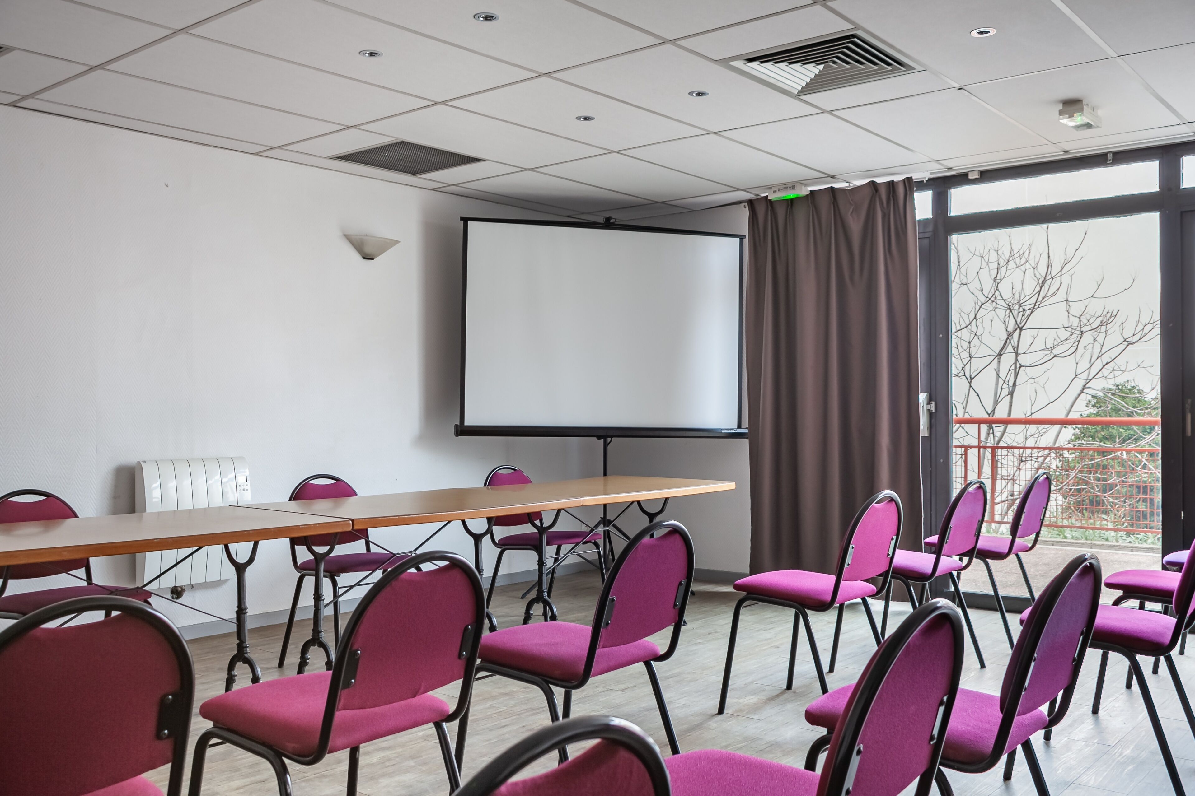 A conference room with a large table, multiple chairs, a projector screen, and a view through a large window.