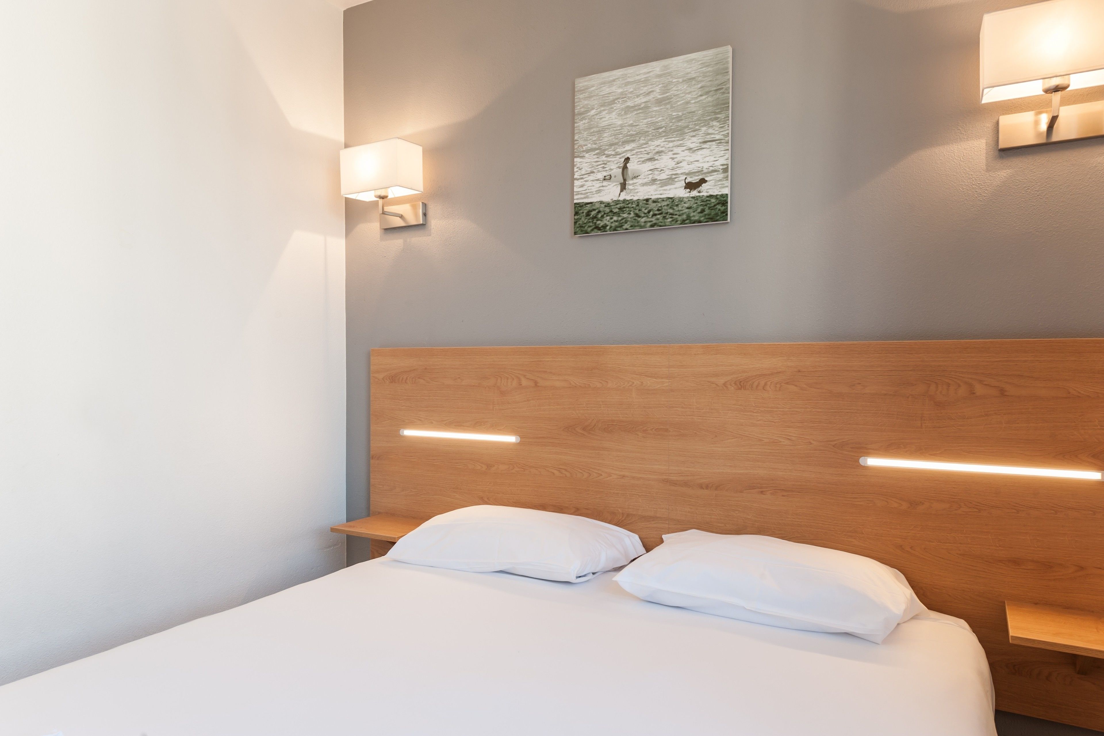 A hotel room with a wooden headboard, two bedside tables, white bedding, and wall-mounted lighting.