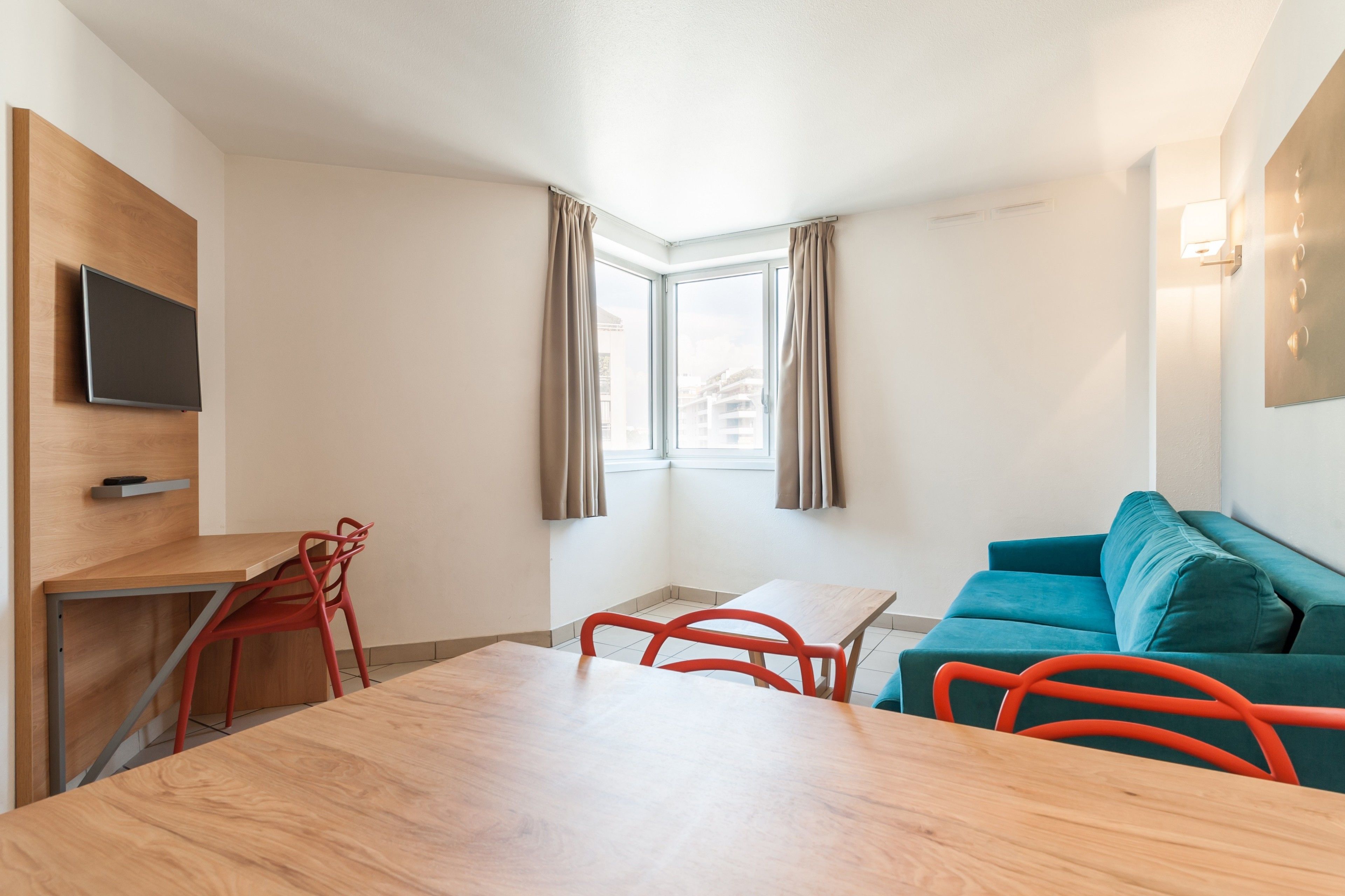 A modern room with a wooden desk, red chairs, a teal sofa, a flat-screen TV mounted on a wooden panel, and a window with curtains.