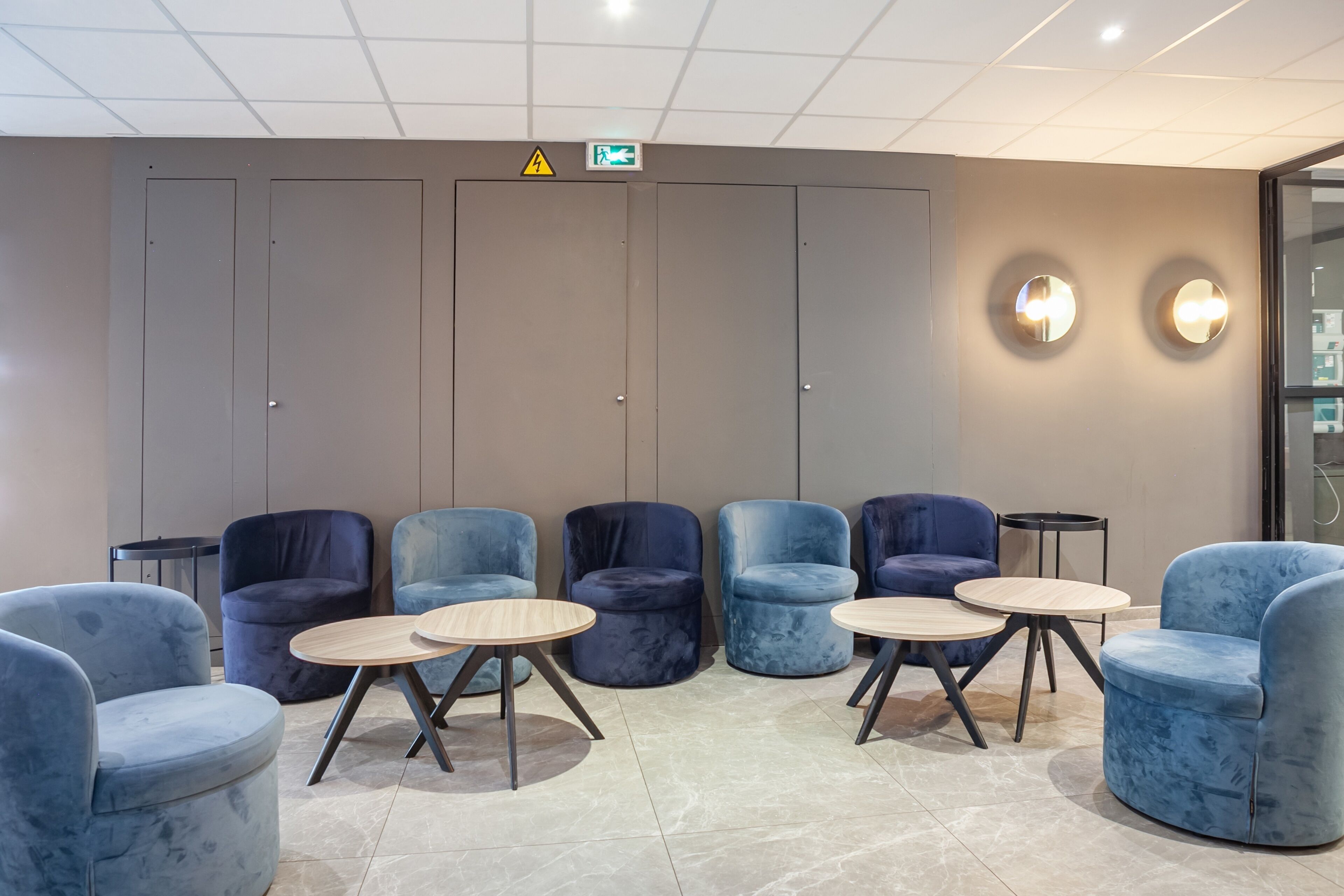 Une salle d’attente avec des fauteuils bleus et des tables rondes, un plafond doté d’un éclairage encastré et un panneau indiquant la sortie de secours, fixé au mur.