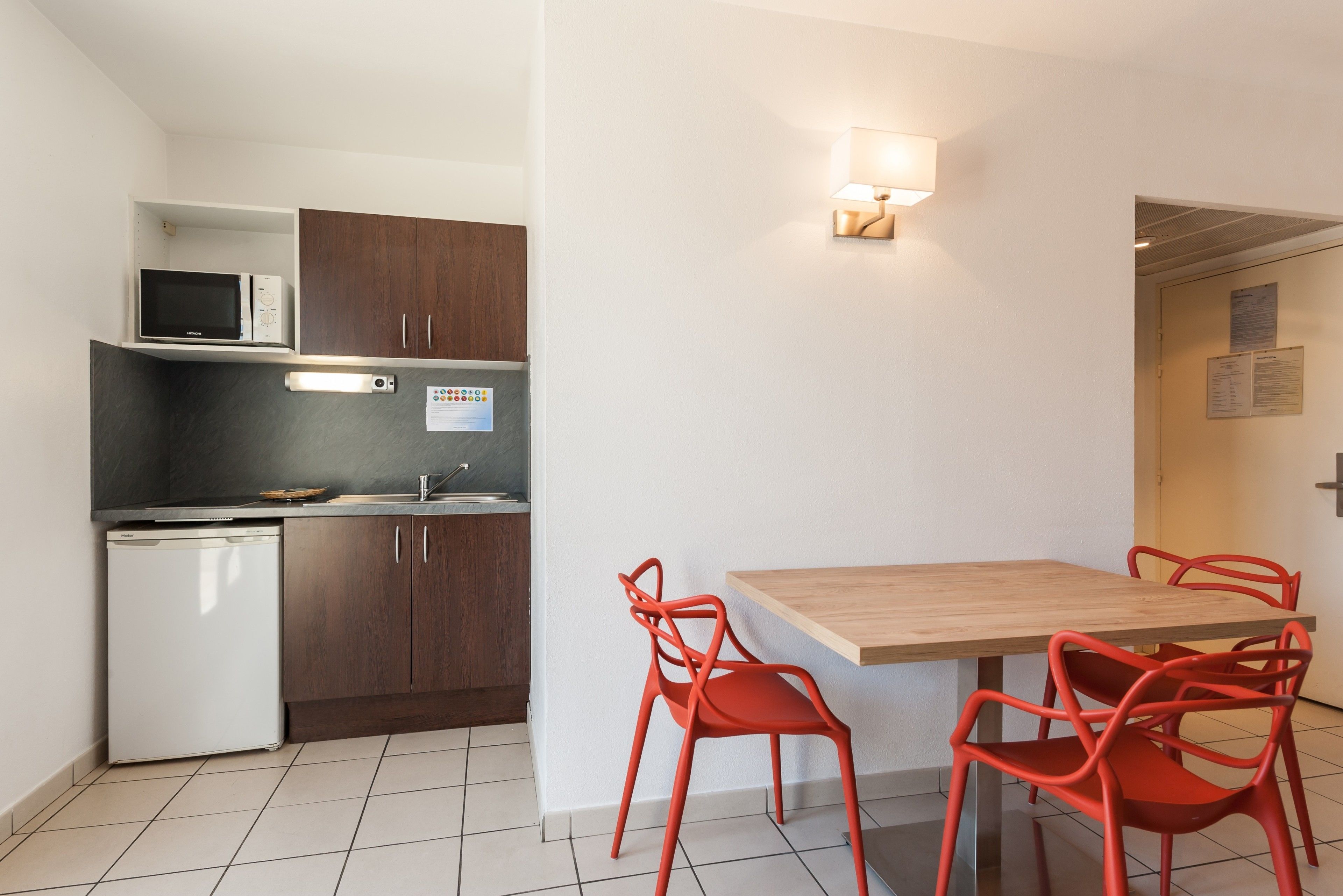 A compact kitchen with a microwave, refrigerator, and wooden cabinets. There is a dining table with four red chairs and a wall-mounted light fixture.