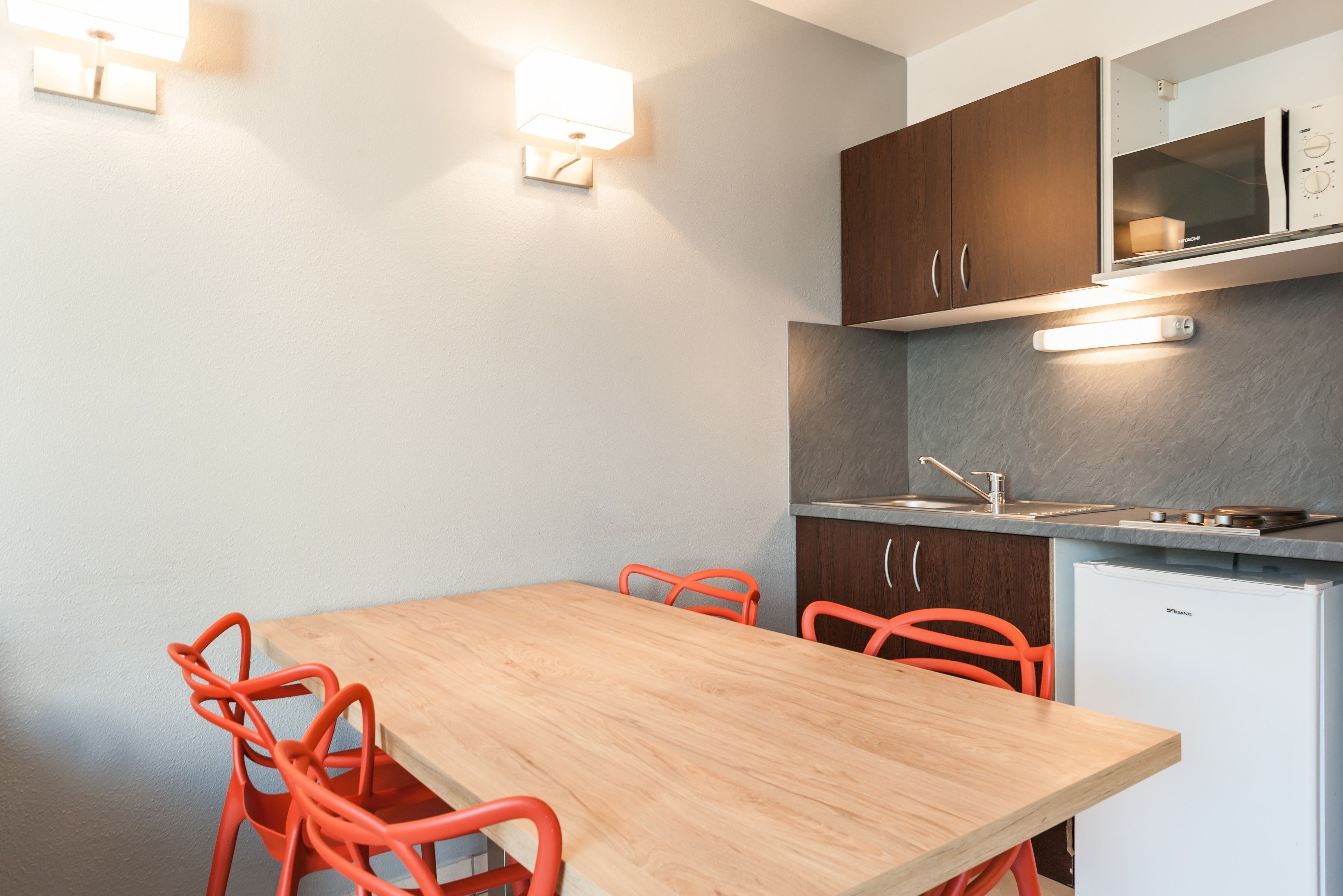 Une cuisine compacte avec une table en bois et des chaises orange, un four encastré et un petit réfrigérateur.
