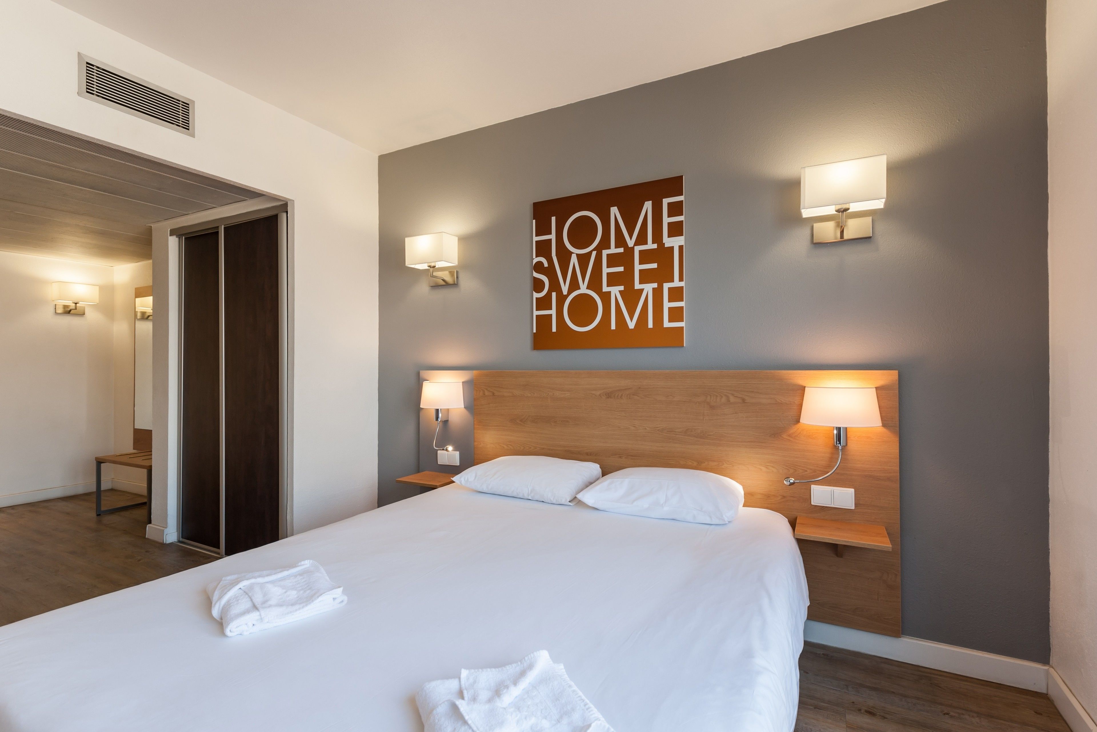 A hotel room with a large bed, two bedside tables with lamps, a wooden headboard, and a wall-mounted sign that reads "HOME SWEET HOME".