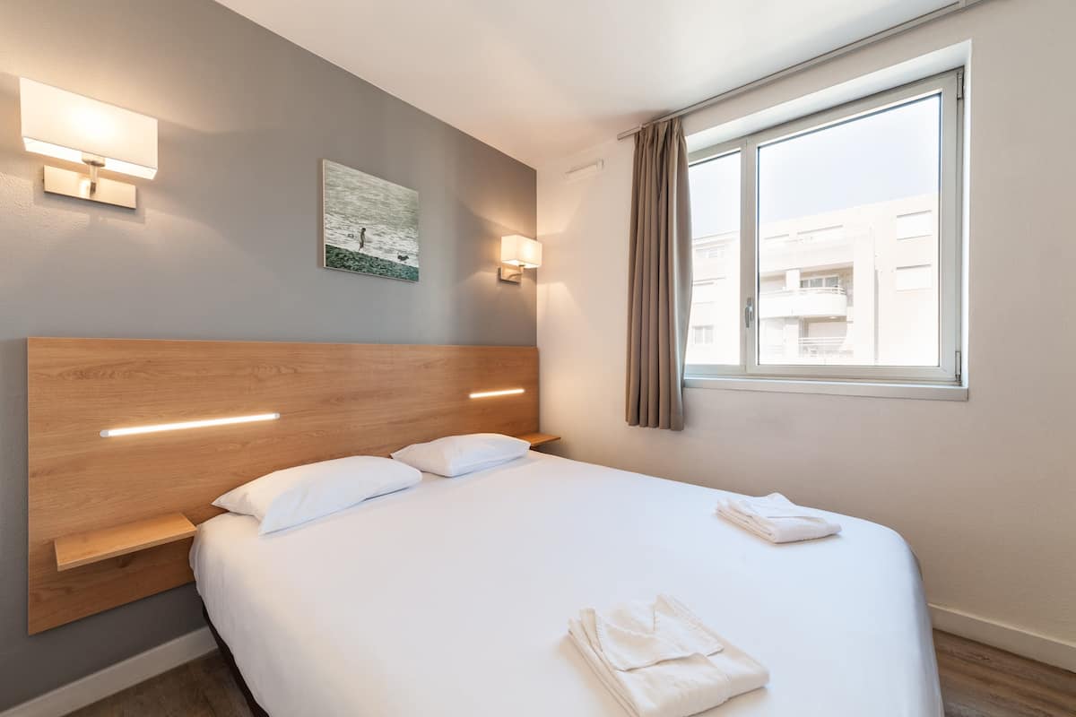 Ein modernes Hotelzimmer mit einem Holzkopfteil, einem großen Bett, zwei Wandleuchten, einem Fenster mit Vorhängen und einem Bild an der Wand.
