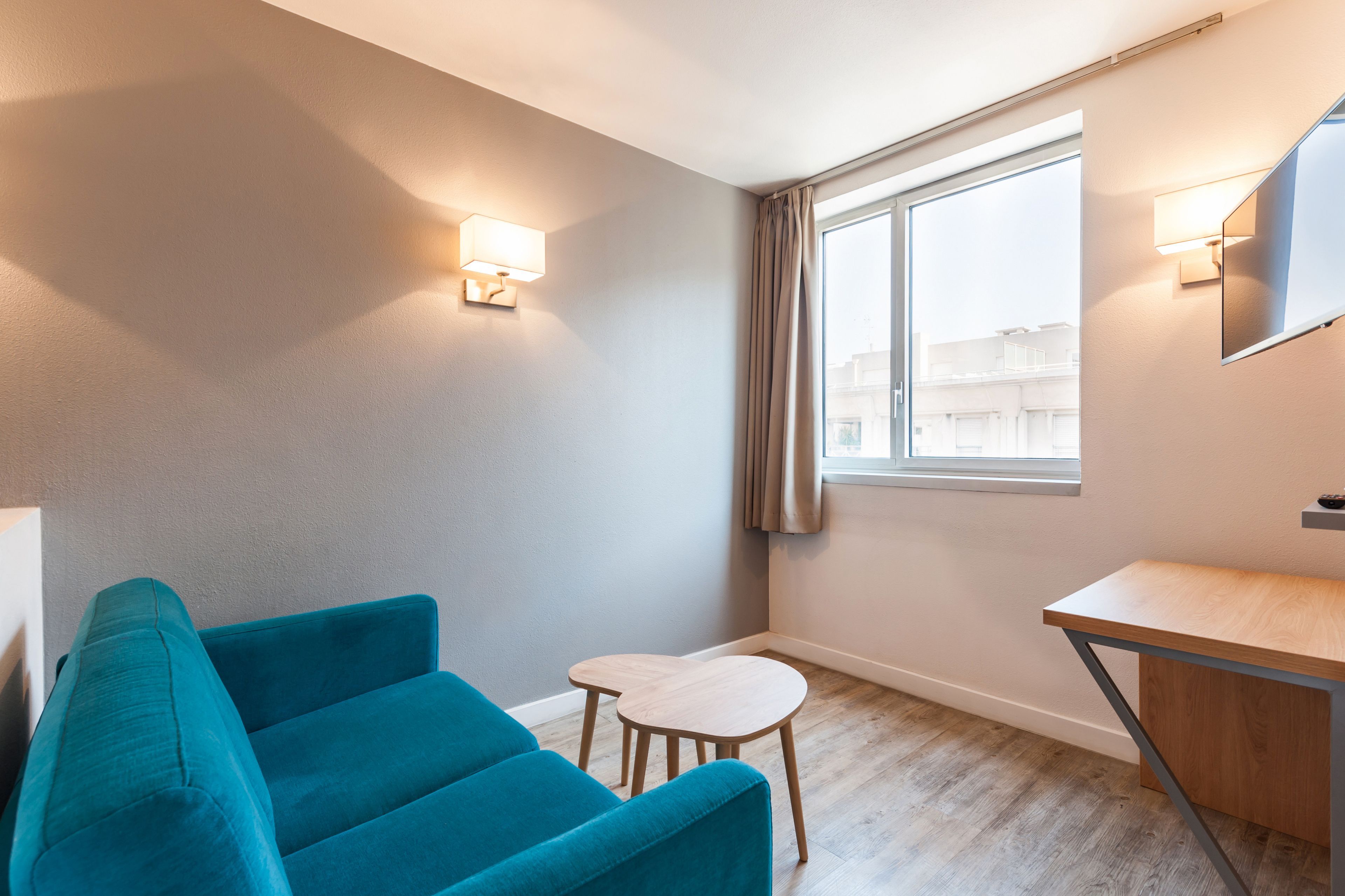 Ein modernes Hotelzimmer mit einem türkisfarbenen Sofa, einem kleinen Holztisch und einem Schreibtisch mit Spiegel.