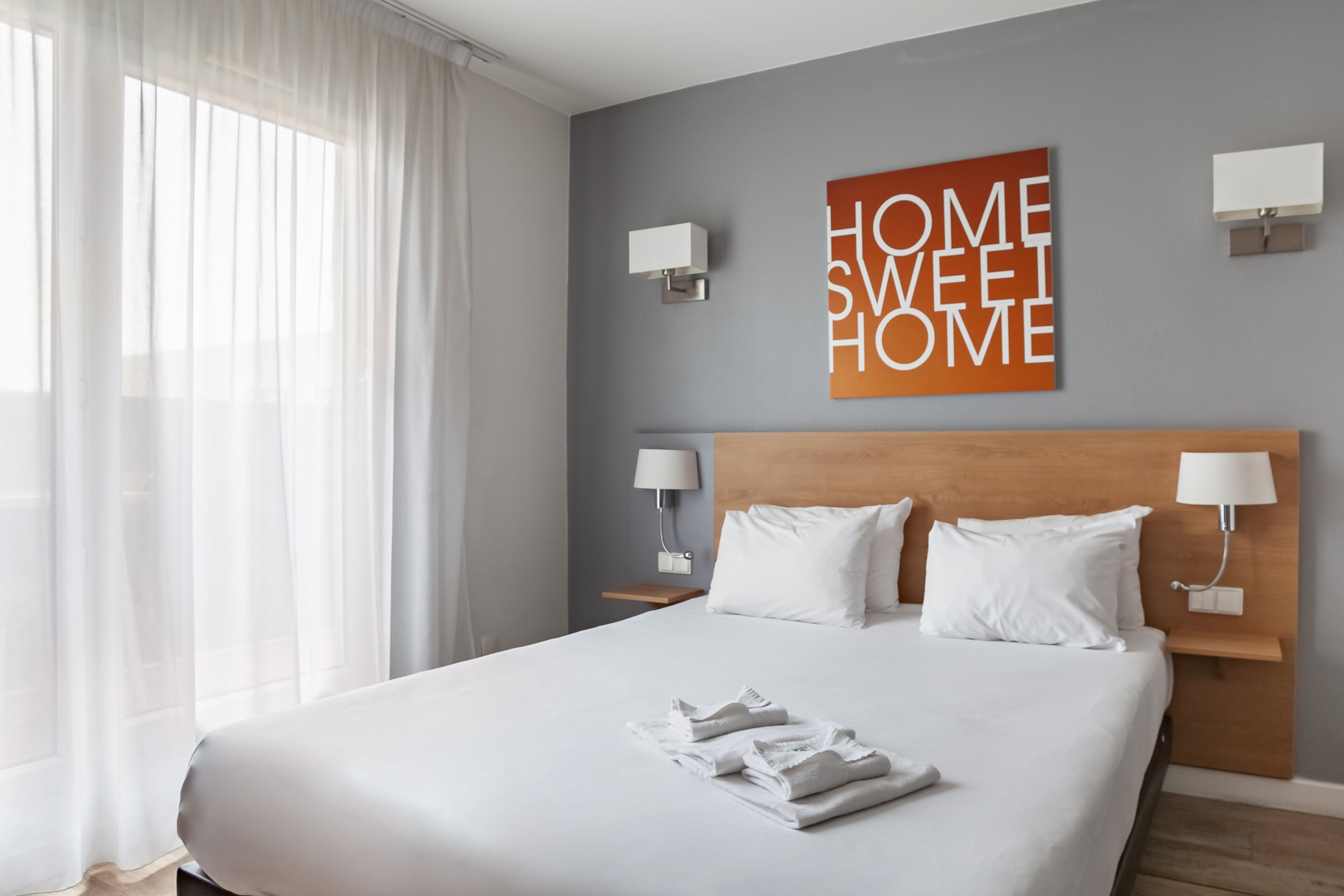 A hotel room with a large bed, white linens, two bedside tables with lamps, a grey wall with a 'HOME SWEET HOME' sign, and sheer curtains.
