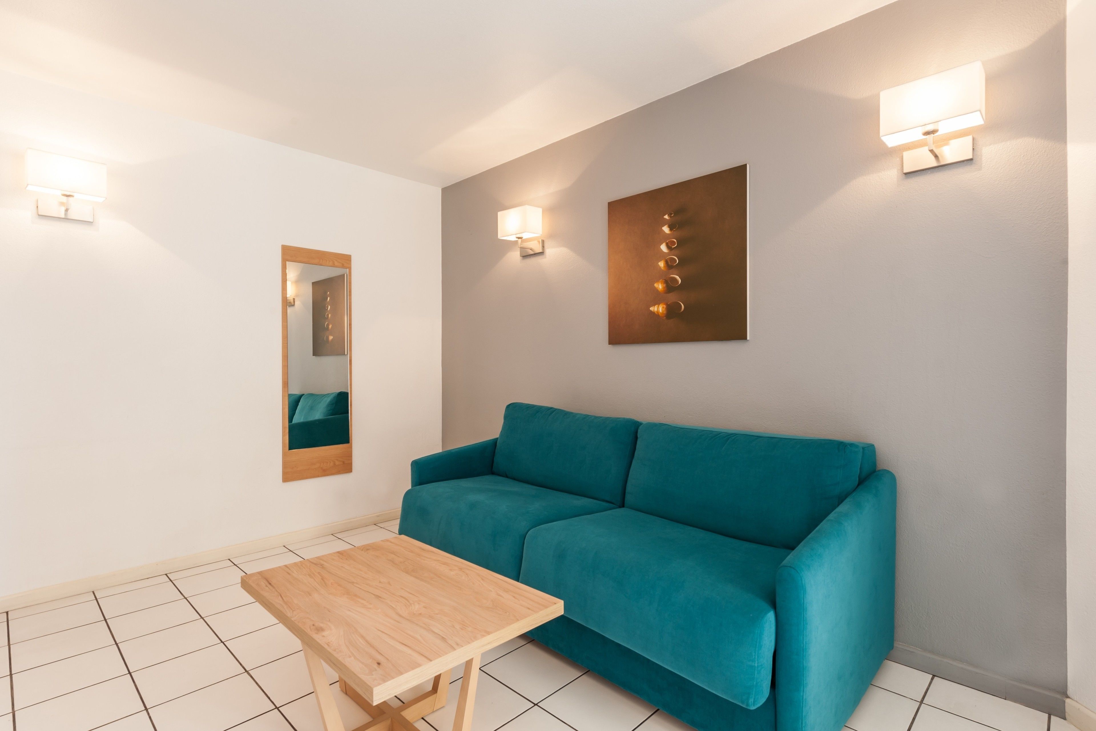 A room with a teal sofa, a wooden table, a mirror, and wall-mounted lights.