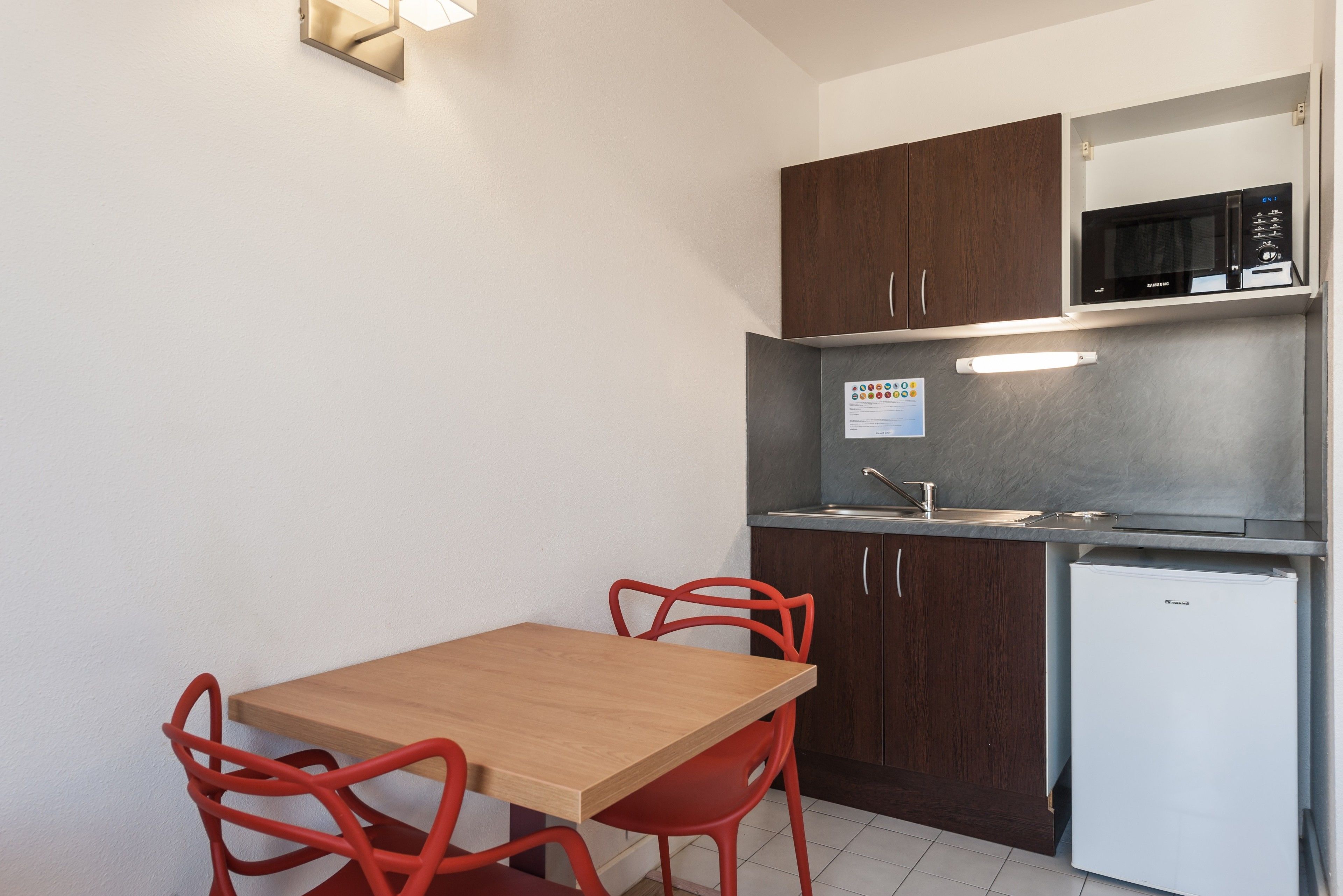 A compact kitchen with dark wooden cabinets, a stainless steel microwave, and a white refrigerator. There is a wooden table with red chairs and a light fixture above it.
