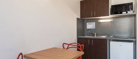 A compact kitchen with dark wooden cabinets, a stainless steel microwave, and a white refrigerator. There is a wooden table with red chairs and a light fixture above it.