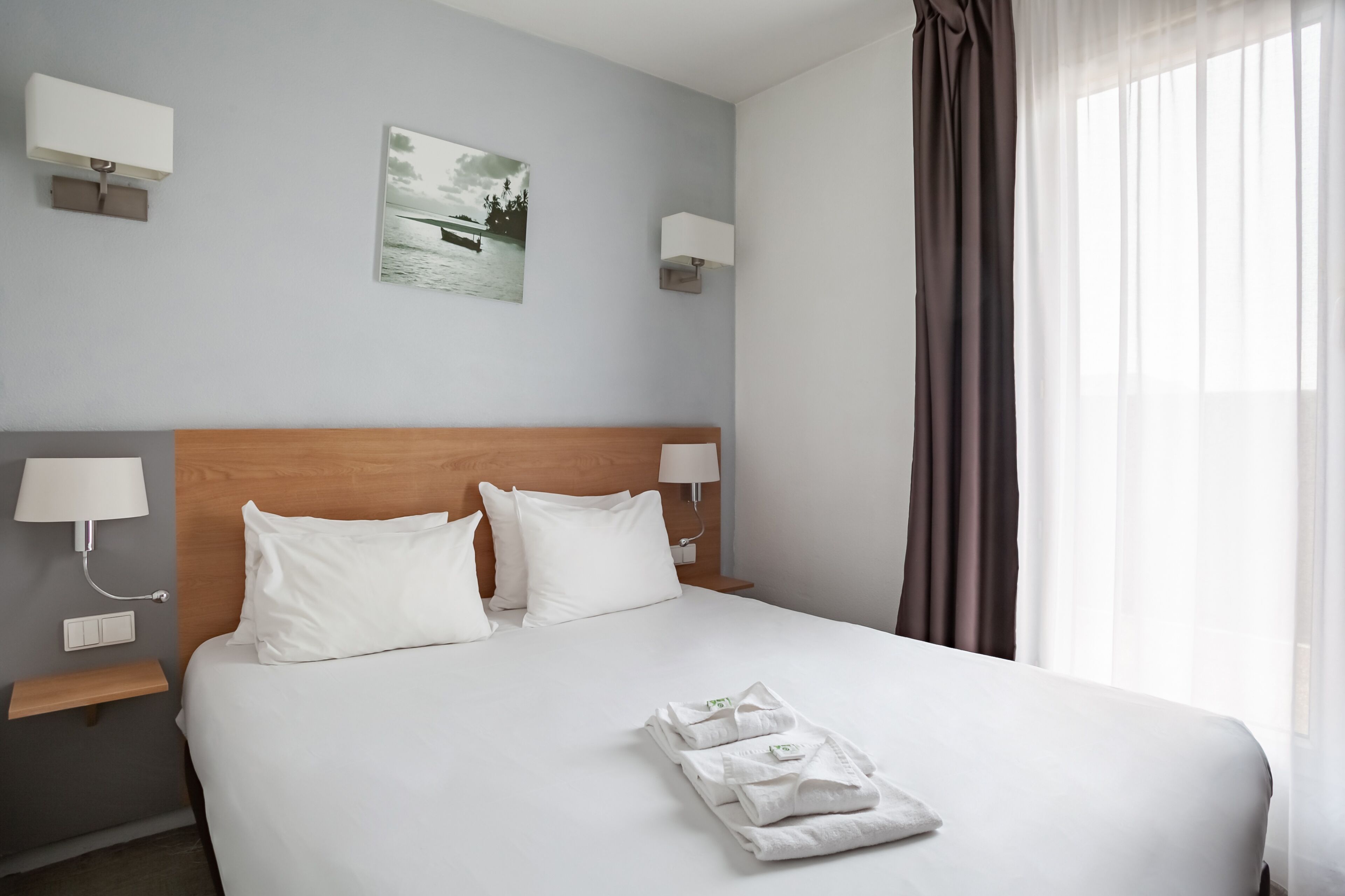A hotel room with a large bed, two bedside tables with lamps, a painting on the wall, and a window with curtains.