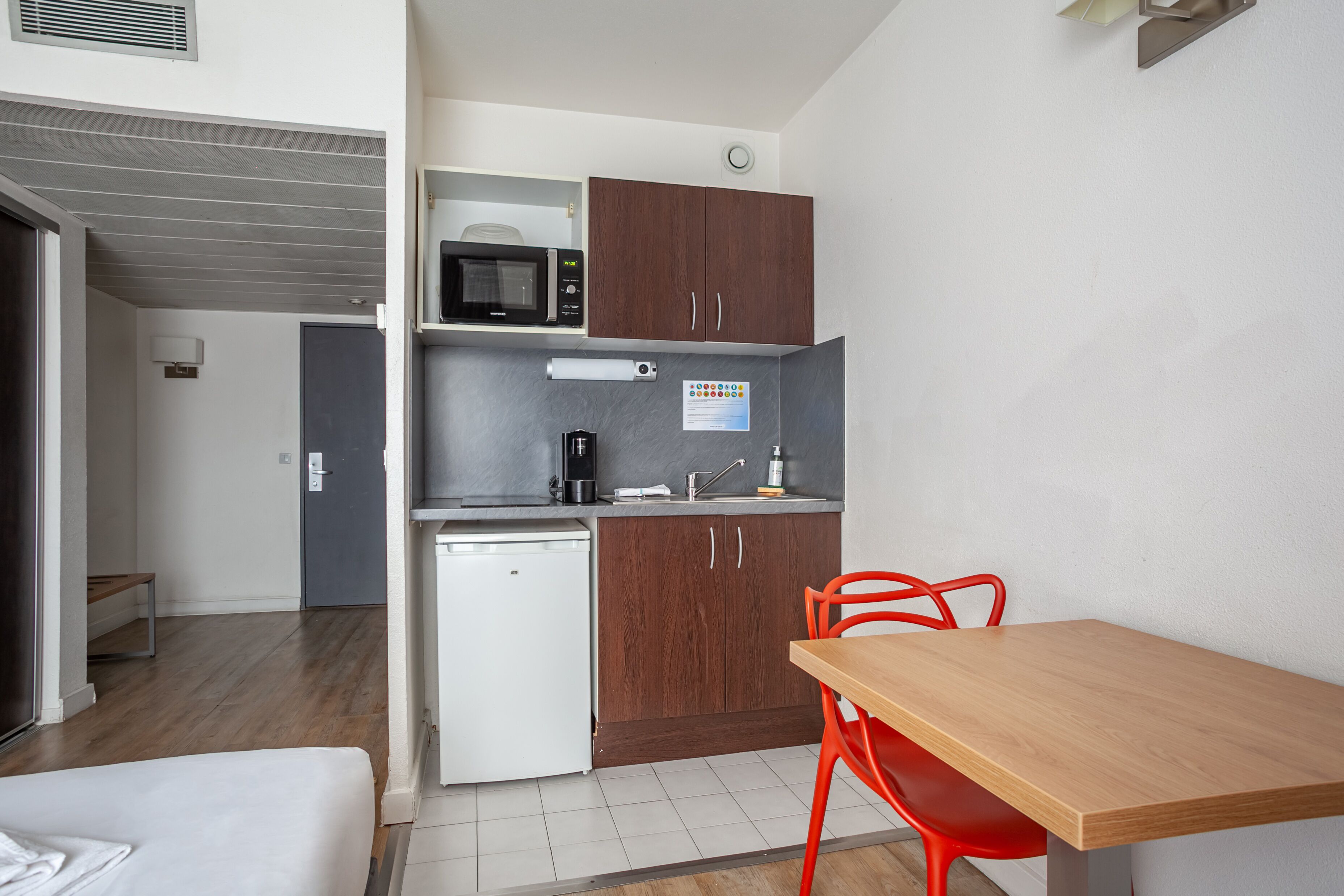 Una kitchenette compacta con microondas, cafetera y lavabo. Cuenta con un refrigerador pequeño, una mesa de madera y una silla roja.