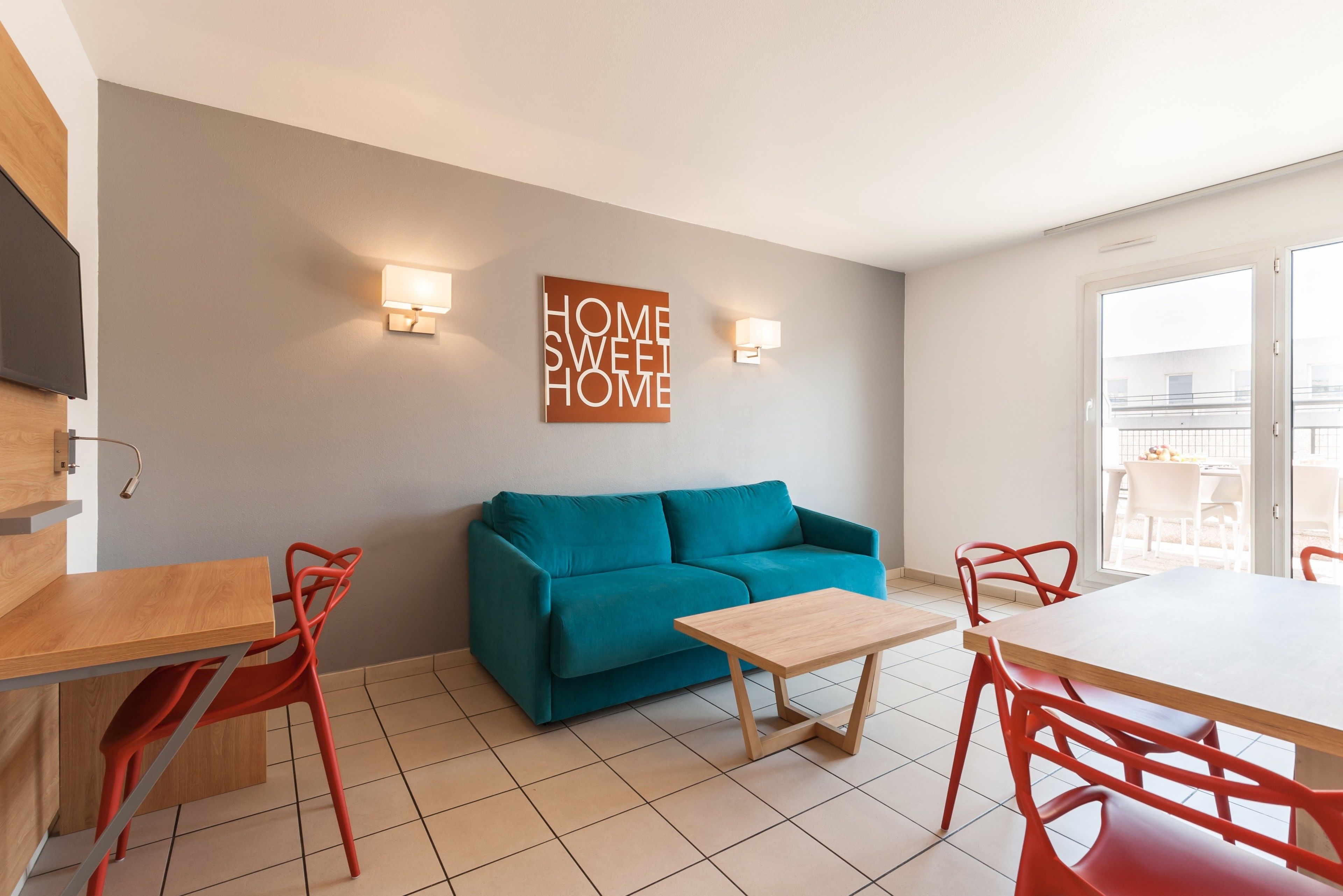 A modern living room with a teal sofa, wooden coffee table, red chairs, and a wall-mounted TV.