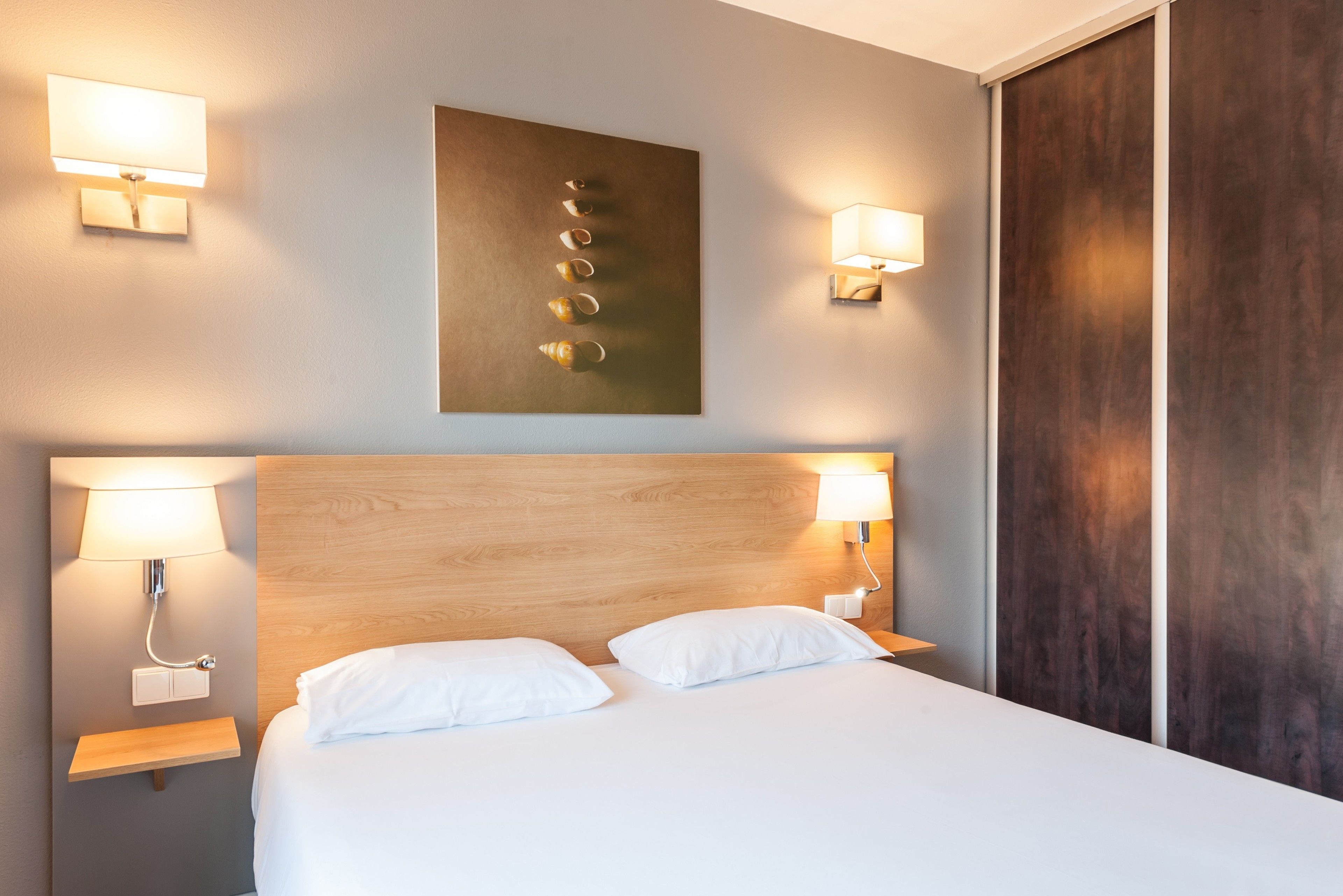 A hotel room with a wooden headboard, two bedside lamps, a wall-mounted artwork, and a large bed with white bedding.