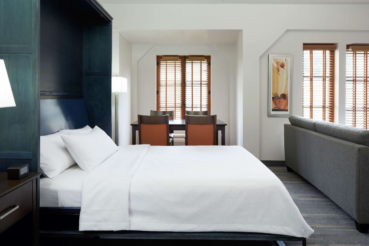 A hotel room with a bed, a desk with chairs, a sofa, and large windows with blinds.
