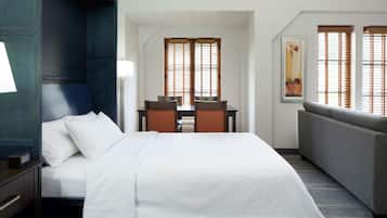 A hotel room with a bed, a desk with chairs, a sofa, and large windows with blinds.
