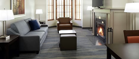 A hotel room with a sofa, armchair, ottoman, flat-screen TV, fireplace, and large windows.