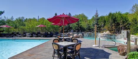 Zona junto a la piscina con mesa y sillas, piscina y una valla con árboles al fondo.