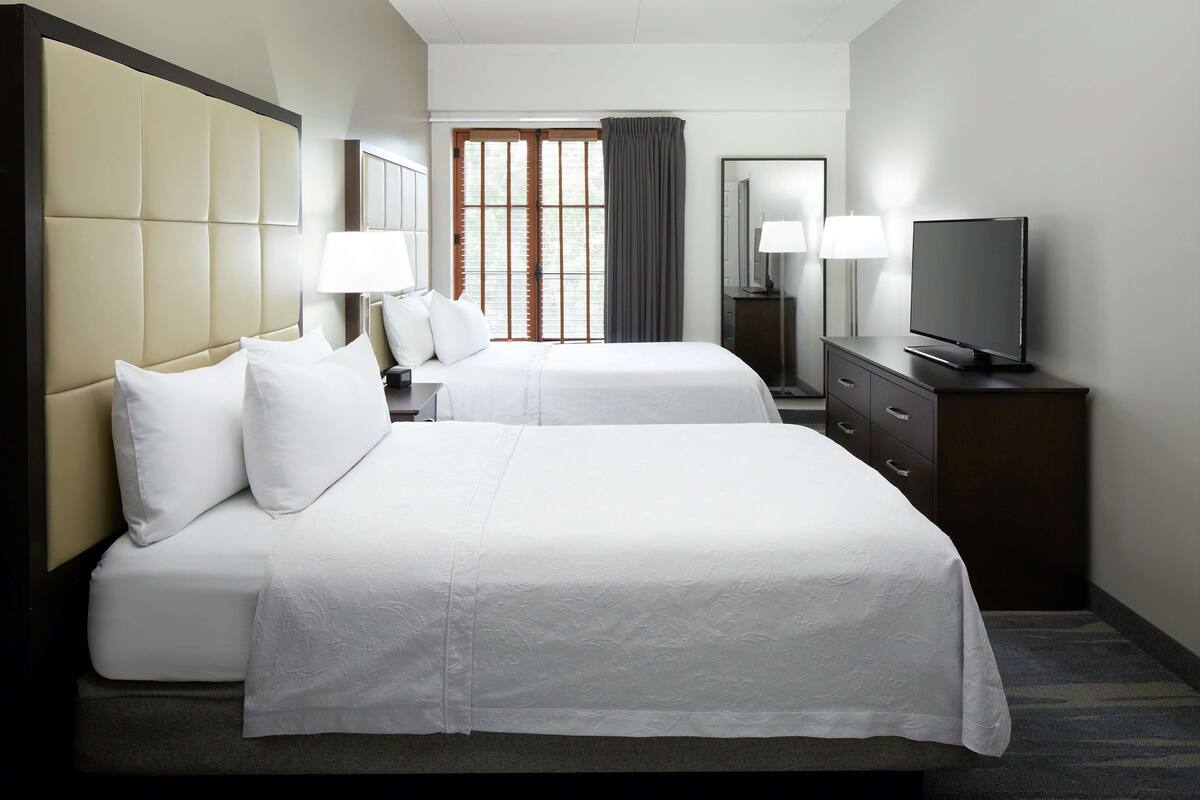 A hotel room with two beds, a TV, a dresser, and a window with curtains.