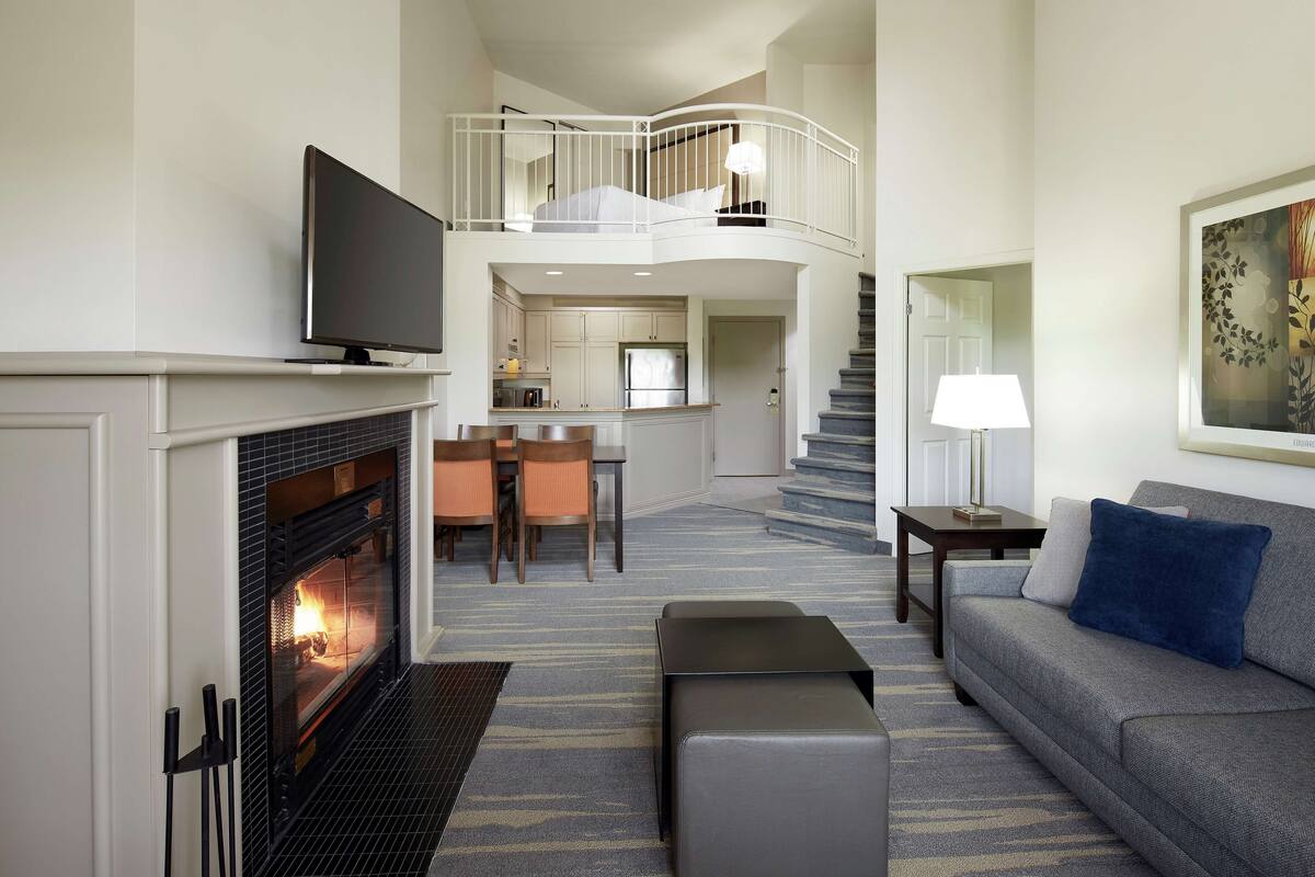 A modern living room with a fireplace, a sofa, a flat-screen TV, and a staircase leading to an upper level.