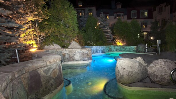 A nighttime swimming pool with clear water, surrounded by rocks and trees, and illuminated by artificial lighting.