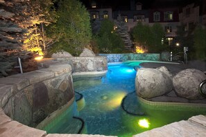 A nighttime swimming pool with clear water, surrounded by rocks and trees, and illuminated by artificial lighting. - Homewood Suites by Hilton Mont-Tremblant Resort (Mont-Tremblant)