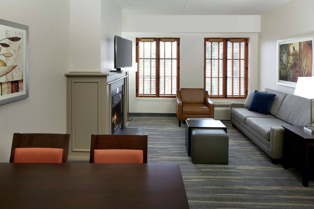 A hotel room with a dining table, two chairs, a sofa, a TV, and two windows with blinds.
