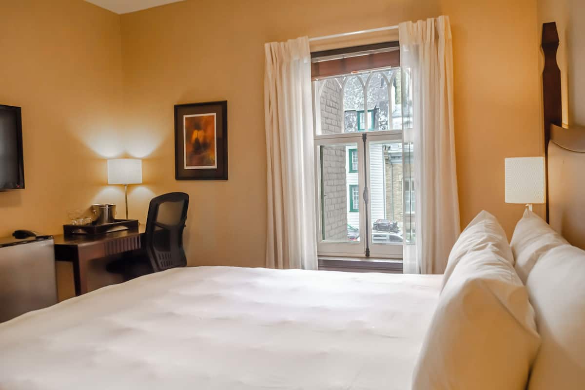 Una habitación de hotel con una cama grande, un escritorio con lámpara, una silla y un ventanal con cortinas.