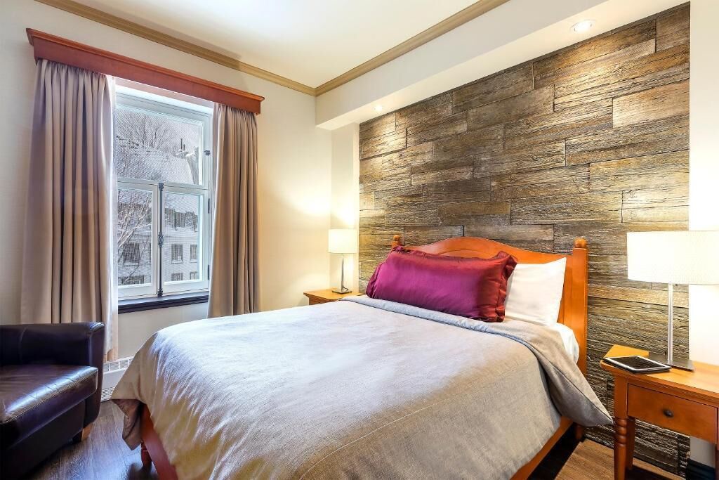 A hotel room with a large bed, a wooden headboard, a nightstand with a lamp, a window with curtains, and a view of buildings outside.